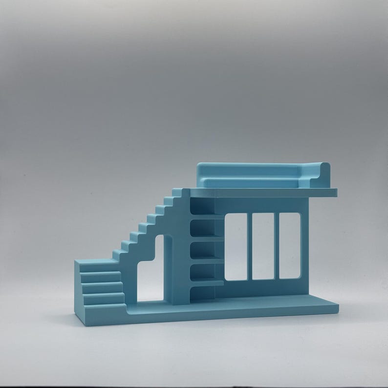 May include: A light blue architectural model featuring stairs, shelves, and a window-like aperture. The model has a flat base and a rectangular top. The design is minimalist and geometric, with clean lines and a solid colour.