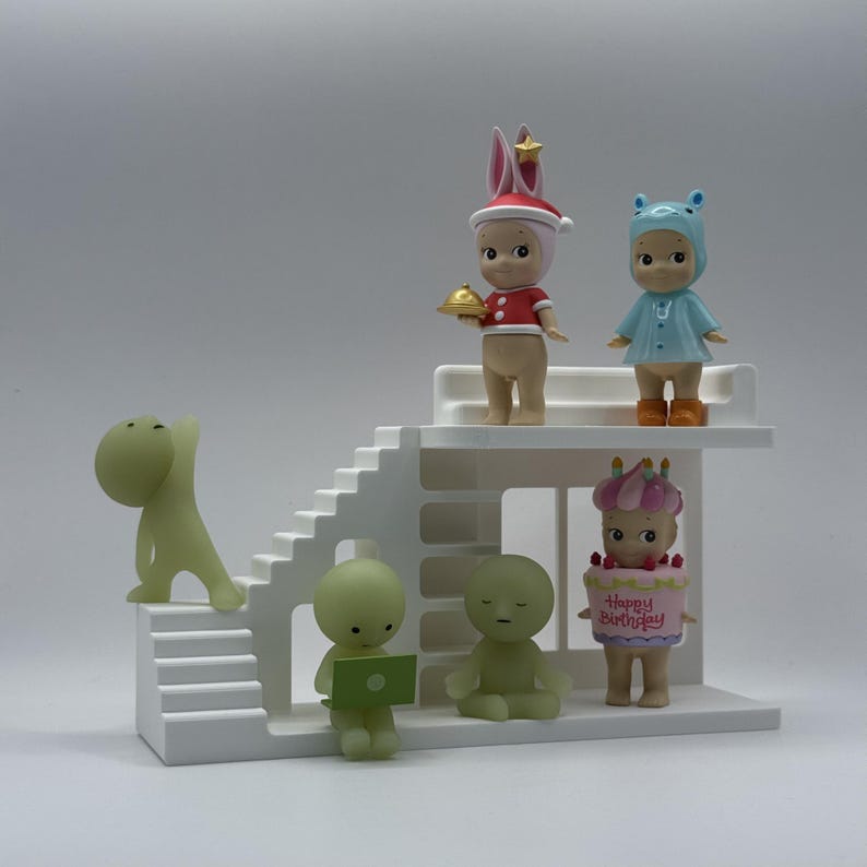 May include: A collection of small, stylized figurines in various poses and outfits, including a bunny, a bear, and a figure holding a laptop. The figurines are arranged on a white, multi-level platform with stairs.