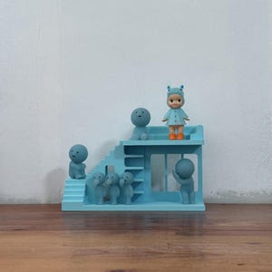 May include: A light blue miniature playset with a staircase and a platform, populated by small, stylized figures. A doll in a blue outfit and orange wellington boots stands on the platform. The figures are various sizes and poses, creating a whimsical scene.