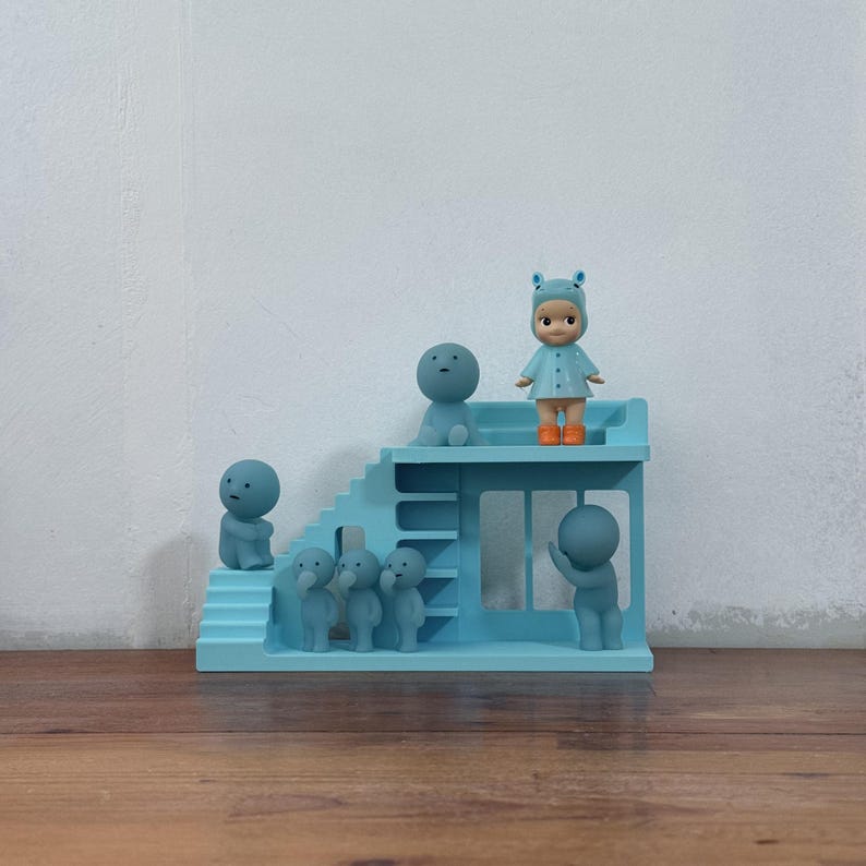 May include: A light blue miniature playset with a staircase and a platform, populated by small, stylized figures. A doll in a blue outfit and orange wellington boots stands on the platform. The figures are various sizes and poses, creating a whimsical scene.