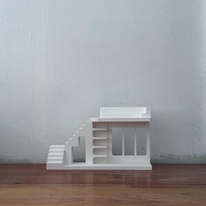 May include: A white miniature architectural model of a building with stairs, a balcony, and a storefront design. The model is made of a solid white material and sits on a wooden surface against a white wall.