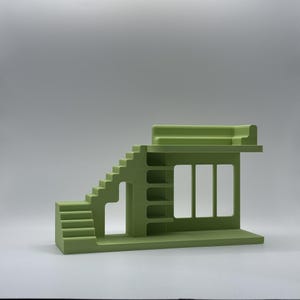 May include: A light green architectural model featuring stairs, a window, and shelves. The model has a flat roof and a rectangular base. The design is minimalist and geometric, with clean lines and a solid colour.