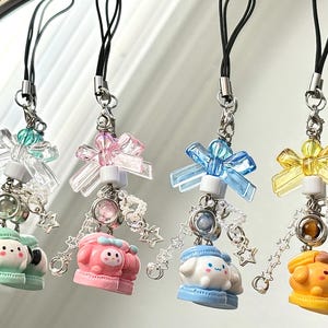 May include: Four colorful phone charms with different cartoon characters. Each charm features a bow, a crystal, and a chain with star-shaped beads.