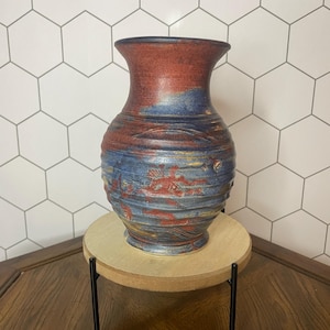 May include: A blue and red ceramic vase with a textured surface. The vase is sitting on a wooden stand with black metal legs.