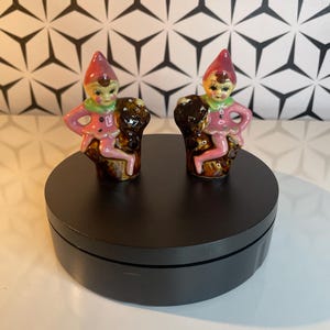 May include: Two ceramic elf figurines. Each elf has a pink outfit, a red hat, and a green collar. They are perched on a brown base. The background is a black and white geometric pattern. The figurines are on a black rotating display stand.
