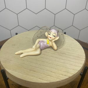 May include: A ceramic figurine of a reclining fairy with a lavender swimsuit and yellow accent. The fairy is resting on a clear, scalloped-edged base. The figurine is displayed on a small, round wooden table with black legs.