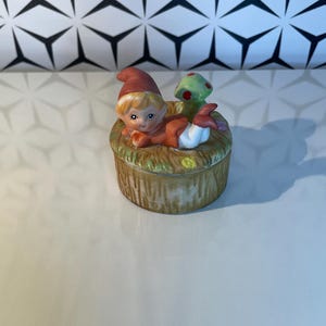May include: A ceramic figurine of a small elf wearing an orange outfit and a red hat, lying on a round, brown base with a green mushroom. The figurine has blonde hair and a smiling face. The base has a textured, wood-like appearance.