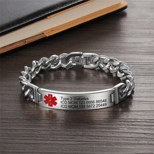 Può includere: Un braccialetto medico identificativo color argento con un design a catena. La placca rettangolare presenta il simbolo rosso della Stella della Vita e il testo: "Type 2 Diabetes" e informazioni mediche. Il braccialetto è su una superficie di legno.
