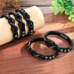 Op de afbeelding: Zwarte gepersonaliseerde armbanden met namen en de zin "Always and forever". De armbanden worden gepresenteerd op een houten oppervlak en een beige cilinder. De armbanden hebben een uniek infinity symbool ontwerp.