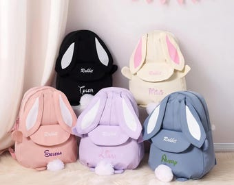 May include: Five colorful bunny-shaped backpacks. Each backpack features large, floppy ears, a fluffy tail, and the word "Rabbit" embroidered on the flap. The backpacks are in black, cream, pink, lavender, and blue, with personalized names embroidered.