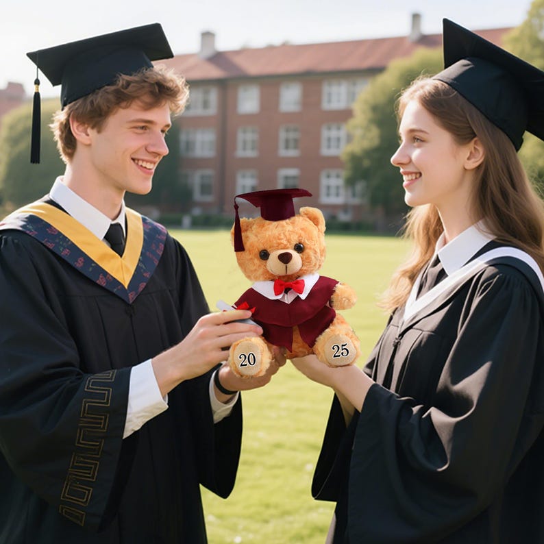 May include: A teddy bear wearing a graduation gown and cap, held by two people in graduation attire. The bear has a maroon gown and cap, a red bow tie, and the numbers "20" and "25" are printed on its paws. The background shows a building and greenery.