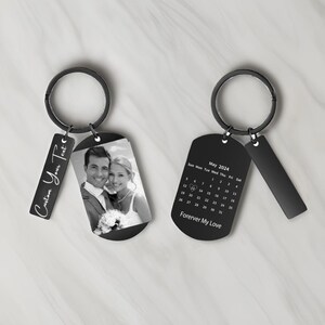 以下が含まれることがあります： 2つの黒いキーホルダー。1つはカップルの写真、もう1つは「Forever My Love」と書かれた2024年5月のカレンダーです。どちらにもテキストの小さなタグが付いています。1つは「Custom Your Text」と書かれています。