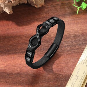 Op de afbeelding: Zwarte leren armband met een uniek infinity symbool ontwerp. De namen "Daine", "Tania", "Kane" en "Olivia" zijn gegraveerd op de band. De woorden "Always and forever" zijn ook zichtbaar. Met magnetische sluiting.