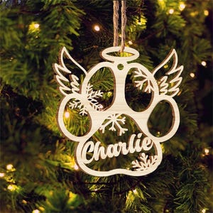May include: A wooden paw print Christmas ornament with angel wings and the name "Charlie" inscribed on it. The ornament is decorated with snowflakes and has a halo above it.
