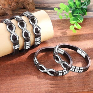 Op de afbeelding: Een verzameling gepersonaliseerde armbanden met donkerbruine leren bandjes en zilverkleurige oneindigheidssymbolen. Elke armband heeft gegraveerde namen en de tekst "Always and forever."