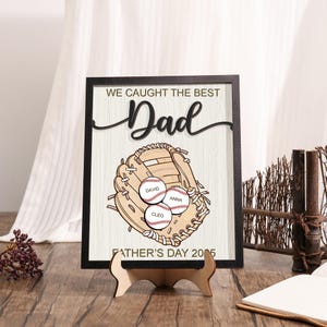 Puede incluir: Un regalo enmarcado para el Día del Padre con un guante de béisbol y tres pelotas de béisbol, cada una con un nombre. El texto dice "WE CAUGHT THE BEST Dad" encima del guante y "FATHER'S DAY 2005" debajo. El marco es negro.
