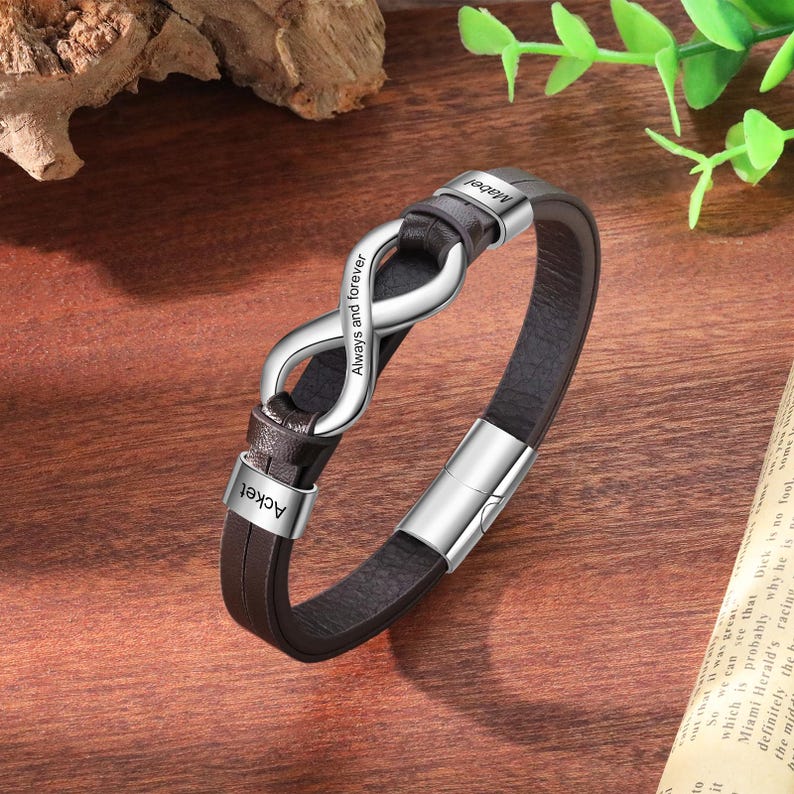 Op de afbeelding: Een bruin leren armband met een zilveren oneindigheidssymbool in het midden. Op het oneindigheidssymbool staat de tekst "Always and forever". De namen "Acket" en "Mabel" zijn ook op de armband gegraveerd.