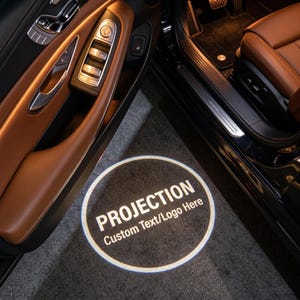 May include: A car door puddle light projector casting a circular white light onto the ground. The projection displays the word PROJECTION in large capital letters above the smaller text Custom Text/Logo Here, framed by a thin circular border.