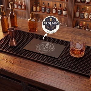 May include: A rectangular black rubber bar mat with a textured nub surface, featuring a central custom logo print that reads THE ALE HOUSE with a beer glass graphic. The mat is placed on a wooden bar top next to a copper jigger and a glass of whiskey.