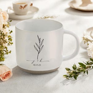 Pode incluir: Caneca de cerâmica branca com um design minimalista. A caneca apresenta uma ilustração botânica preta e a inscrição "j + h" com a data "02.15.25" em uma escrita delicada. A base da caneca é de cor cinza neutra.