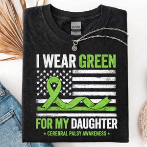 Op de afbeelding: Donkergrijze t-shirt met de tekst "I Wear Green For My Daughter" en een groen lintontwerp over een gestileerde Amerikaanse vlag. De t-shirt is voor Cerebral Palsy Awareness.