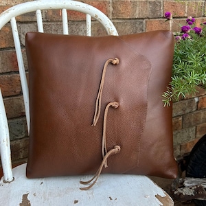May include: A brown leather cushion with a decorative flap and knotted leather ties. The cushion is sitting on a white wooden chair.