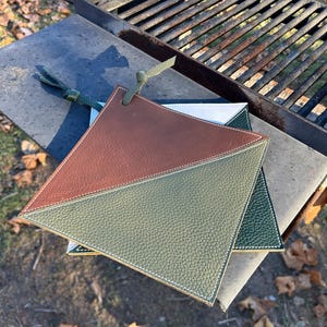 May include: Two leather pot holders in green, brown, and white. The pot holders are triangular in shape and have a stitched edge. They are sitting on a metal grill.