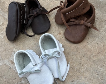 May include: Four pairs of baby moccasins in shades of brown and white. The leather shoes have a scalloped edge and are tied with a bow. The moccasins are arranged on a neutral-colored surface.