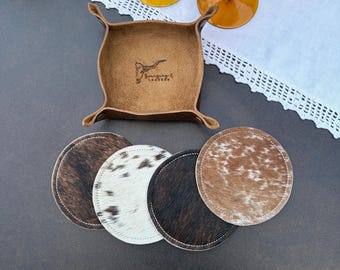 May include: A square tan leather valet tray with snap corners and a stamped logo featuring a longhorn skull, accompanied by four circular coasters with varied brown, white, and black cowhide patterns and stitched edges.