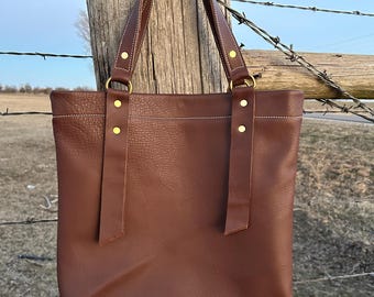 May include: A brown leather tote bag with brass hardware and a white stitch detail. The bag is hanging on a wooden fence post.
