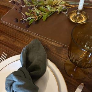 May include: A white plate with a gold rim, a green linen napkin, and a brown leather placemat with a sprig of dried foliage and a brass candle holder with a white candle.