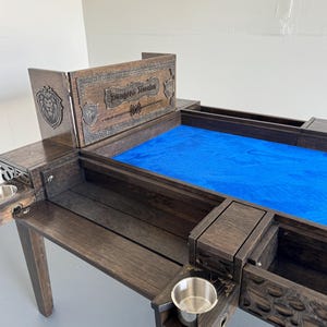 Könnte beinhalten: Dunkel gebeizter Spieltisch aus Holz mit einer blauen Filz-Spielfläche. Der Tisch verfügt über einen "Dungeon Master"-Bildschirm, Getränkehalter und Staufächer. Der Bildschirm hat ein Löwenwappen- und Schilddesign.