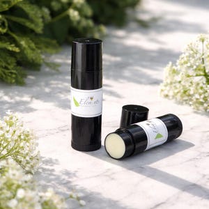 May include: Black lip balm tubes with white labels featuring the word "Elements" and a green leaf design. One tube is open, revealing the balm. The background includes white flowers and green foliage.