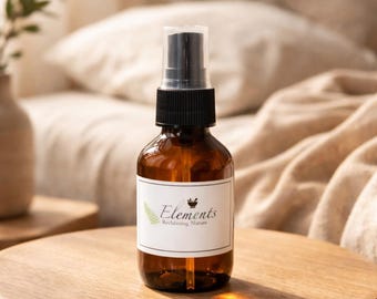 May include: A brown glass spray bottle with a black pump and a white label. The label reads "Elements Reclaiming Nature" with a fern graphic. The bottle is on a wooden surface, with a blurred background of a bed.