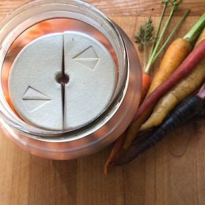 Peut inclure: Un bocal en verre avec un poids en céramique blanche à l'intérieur. Le bocal est rempli de carottes orange et le poids a deux flèches pointant dans des directions opposées. Le bocal est posé sur une surface en bois.