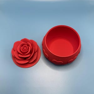 May include: A red, rose-shaped trinket box with a matching lid. The lid is a detailed rose, and the box has a textured design around the sides. The box is open, revealing the interior. The background is a solid light blue.