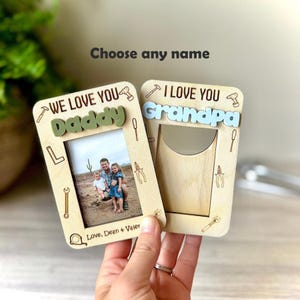 Op de afbeelding: Twee houten fotolijsten met de tekst "WE LOVE YOU Daddy" en "I LOVE YOU Grandpa". De lijsten hebben illustraties van gereedschap en ruimte voor foto's. De tekst "Choose any name" is ook zichtbaar.