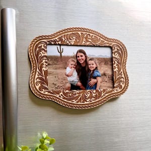 Op de afbeelding: Een rechthoekige houten fotolijst met een decoratieve, lasergesneden rand met een ingewikkeld bloemmotief. De lijst bevat een foto van een volwassene en twee kinderen in een woestijnlandschap met een cactus op de achtergrond.