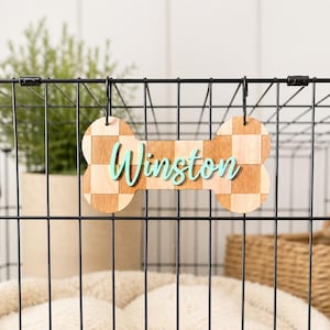 Pode incluir: Uma placa de madeira em forma de osso de cão, com um padrão xadrez e o nome "Winston" em escrita turquesa. A placa está pendurada em uma gaiola de arame preta, com uma planta e uma cesta no fundo.