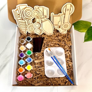 Puede incluir: Un kit de manualidades de madera con un camión de construcción, pinceles, una paleta de pintura y un juego de 12 botes de pintura de varios colores. El kit está empaquetado en una caja blanca con relleno de papel marrón.
