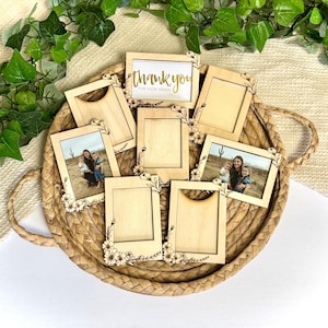 以下が含まれることがあります： 小さな明るい色の木製フォトフレームのコレクション。角には花のアクセントが付いています。一部のフレームには母と子供たちの写真が、1つには「thank you for your order」の文字が金色の文字で表示されています。フレームは編まれたバスケットに配置されています。