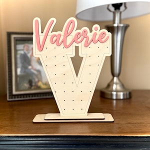 May include: A wooden letter V earring holder with the name "Valerie" in pink lettering on top. The letter V is decorated with small holes for earrings.