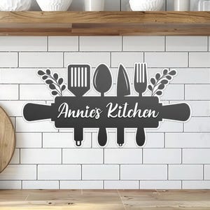 May include: Black and white kitchen sign with the text "Annie's Kitchen" and a rolling pin with a spatula, spoon, knife, and fork on top.