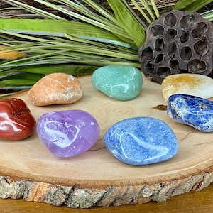 May include: Seven smooth, polished stones in various colors, including red, orange, yellow, green, blue, and purple, arranged in a circle on a wooden surface.