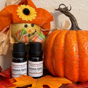 Può includere: Due piccole bottiglie di vetro marrone con tappi neri ed etichette bianche. Le etichette riportano "Mama Zenn Autumn Spice Incense, Clove Bud, Orange, Ginger". Le bottiglie sono appoggiate su un letto di foglie autunnali arancioni e rosse con uno spaventapasseri e una zucca sullo sfondo.