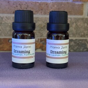 Puede incluir: Dos pequeños frascos de vidrio marrón con tapas negras. Los frascos están etiquetados "Mama Zenn Dreaming" y enumeran los ingredientes como "Naranja, Lavanda, Incienso".