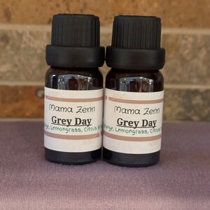 Könnte beinhalten: Zwei kleine braune Glasflaschen mit schwarzen Kappen. Die Flaschen sind mit "Mama Zenn Grey Day" beschriftet und enthalten ätherische Öle. Die Flaschen stehen auf einer violetten, strukturierten Oberfläche.