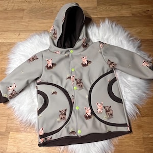 Puede incluir: Chaqueta con capucha gris con interior y puños marrones. La chaqueta presenta un estampado de cerdos y vacas de dibujos animados. Tiene dos bolsillos grandes y curvos con contorno marrón y se abrocha con botones verdes.