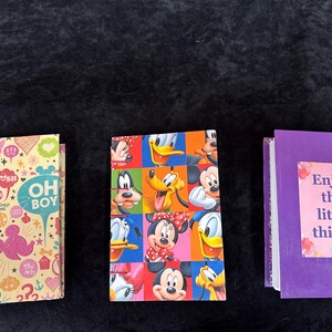 Op de afbeelding: Drie kleine, kleurrijke, gebonden dagboeken. Het eerste dagboek heeft een gele achtergrond met roze, blauwe en groene cartoonfiguren en de tekst "Oh Boy". Het tweede dagboek heeft een veelkleurige achtergrond met cartoonfiguren van Mickey Mouse, Donald Duck en Goofy. Het derde dagboek is paars met de tekst "Geniet van de kleine dingen" op een roze achtergrond.