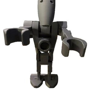 May include: A gray, articulated robot figure with a cylindrical head and two eye-like indentations. The figure has arms with curved hand-like attachments and a circular opening in the torso. The legs are blocky and end in a wide base.